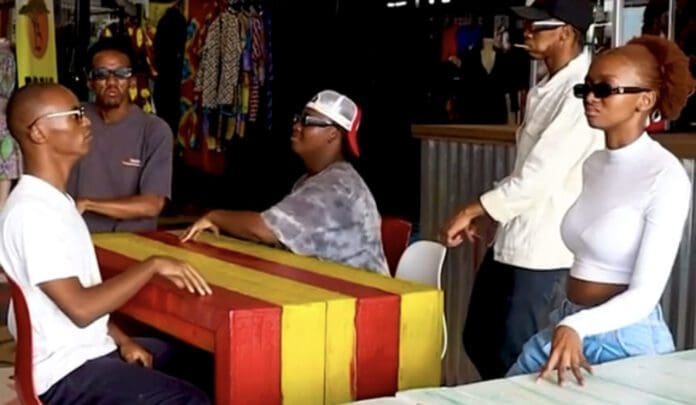 Five people, four seated and one standing, at brightly colored tables in a store with clothing displayed in the background. All are wearing sunglasses and casual clothing, embracing the latest trends inspired by Uyanda Sibiya.