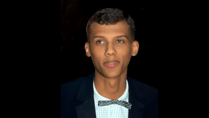 A person with short hair and a bow tie is looking slightly past the camera. He is wearing a dark blazer and a light-colored shirt, dressed as if he's about to perform at a popular music event in 2022.