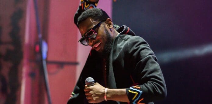 A person wearing sunglasses and a black jacket with colorful accents holds a microphone on stage, smiling and performing music that resonates with the vibrant spirit of Nigeria.