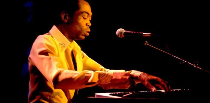 A person wearing a yellow shirt plays the keyboard and sings into a microphone on stage under dim lighting, channeling the spirit of Nigerian icon Fela.