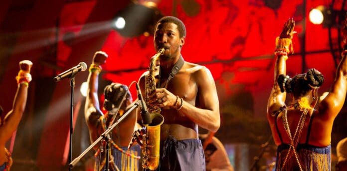 A shirtless man plays a saxophone on stage, surrounded by dancers in colorful attire and accessories with their hands raised. The background, reminiscent of the vibrant energy at the 2022 Grammys, is illuminated with red lighting.