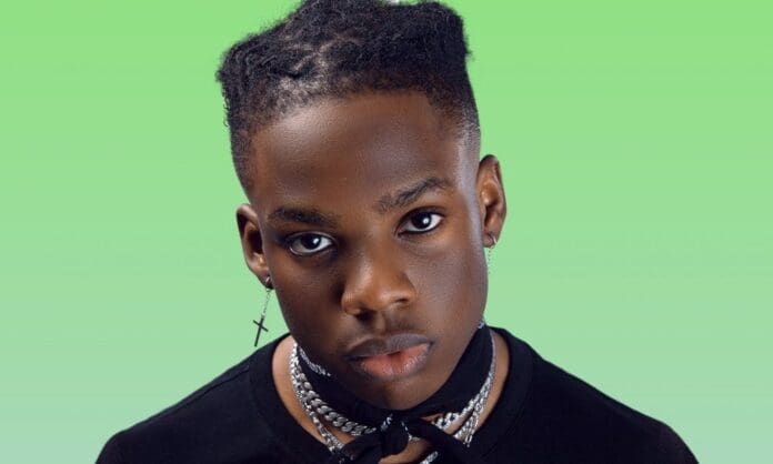 A person with short, stylish hair and wearing a black shirt, multiple silver chains, and an earring shaped like a cross, stands against a green background reminiscent of 2020 music album covers.