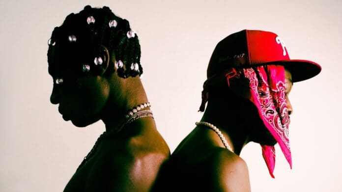 Two individuals are standing back-to-back. One wears bead-adorned braids, while the other sports a red cap and bandana. Both are shirtless and wear pearl necklaces, set against a plain background. Behind the music of Travis & Elzzz, their unique style shines through every detail.