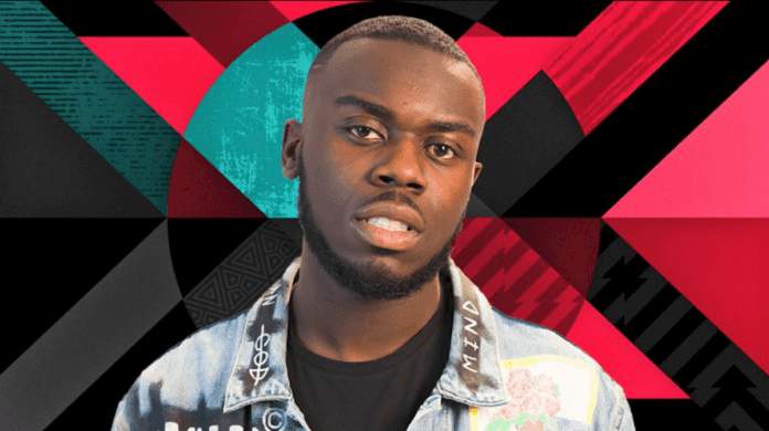 A man with a short beard and mustache stands against a background of geometric shapes in red, black, and teal colors. He is wearing a denim jacket with multiple patches, looking ready for the BBC Radio 1Xtra Rave Show.