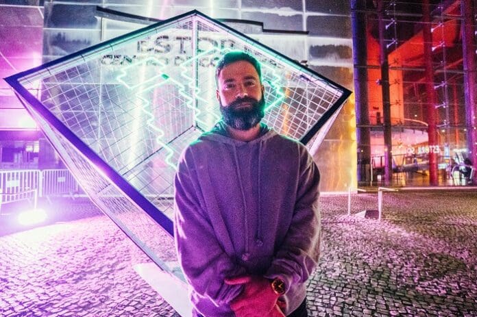 A person with a beard and grey hoodie stands in front of a geometric art installation illuminated by colorful lights, reminiscent of the vibrant atmosphere found at Lisbon afro club.