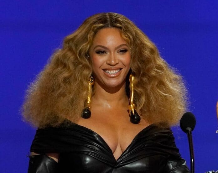 A woman with long, curly hair and wearing a black outfit stands in front of a blue background with a microphone, ready to perform the DJ Kev Karter Remix of Beyonce's 