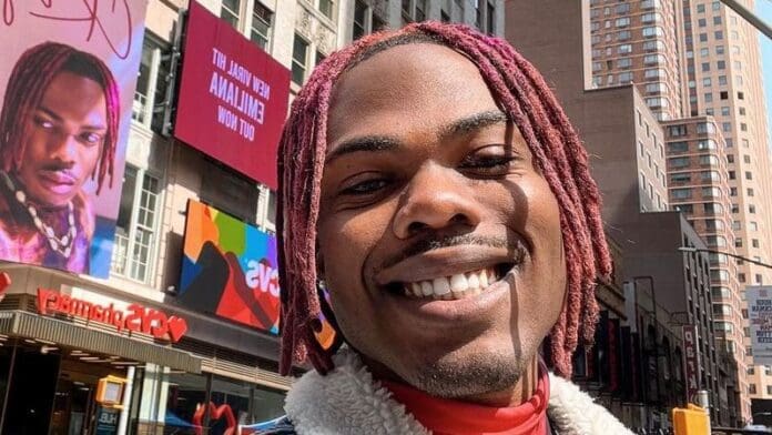 A person with red-dyed hair smiles in an urban setting as a large billboard in the background displays their image with 