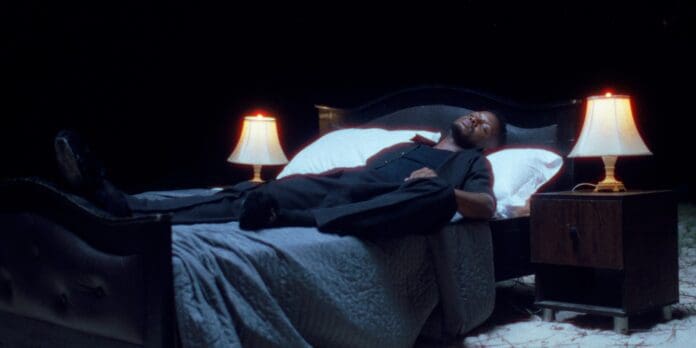 A man lies in bed under a quilt, fully dressed, with two lit lamps on bedside tables on either side, against a black background, as if plucked from an evocative music video by Chike.