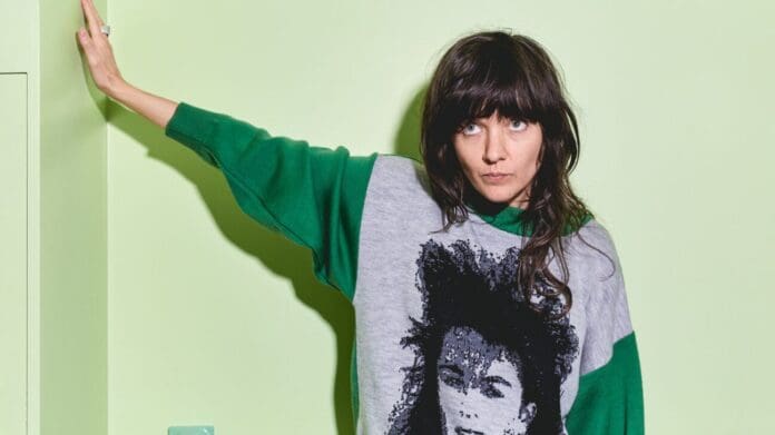 A person with long, dark hair stands against a light green wall, wearing a green sweater with an image of a face on the front. The person, reminiscent of a Marathon Music artist, is looking directly at the camera with one arm raised.