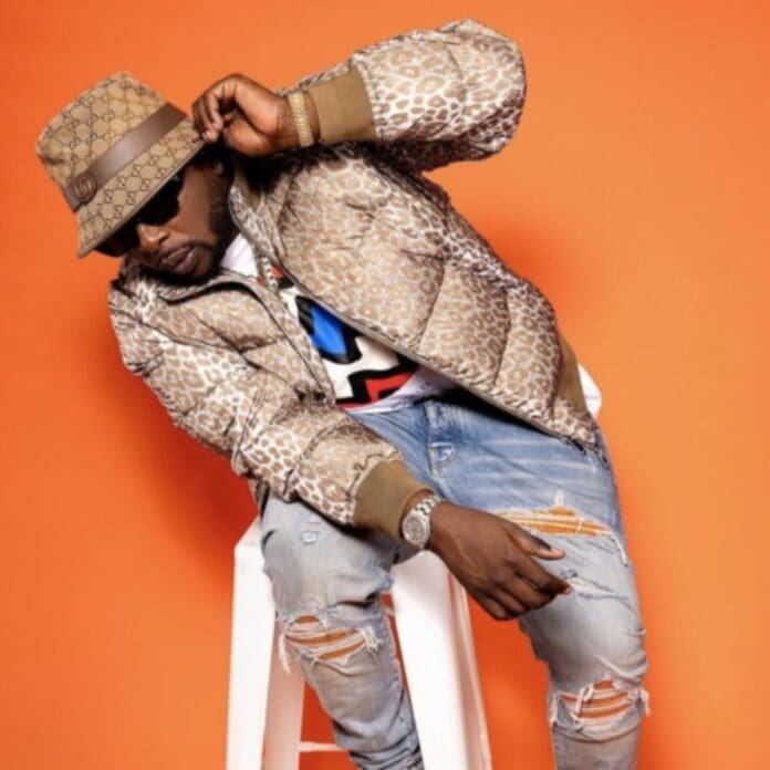 A person wearing a leopard print jacket, a patterned hat, sunglasses, and ripped jeans channels DJ Maphorisa's bold style while posing confidently on a white stool against an orange background.