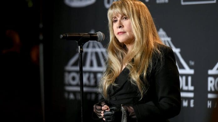 A woman with long blonde hair stands at a microphone on a stage with Rock & Roll Hall of Fame logos in the background. Dressed in a black outfit and gloves, she channels the iconic Stevie Nicks with her commanding presence.