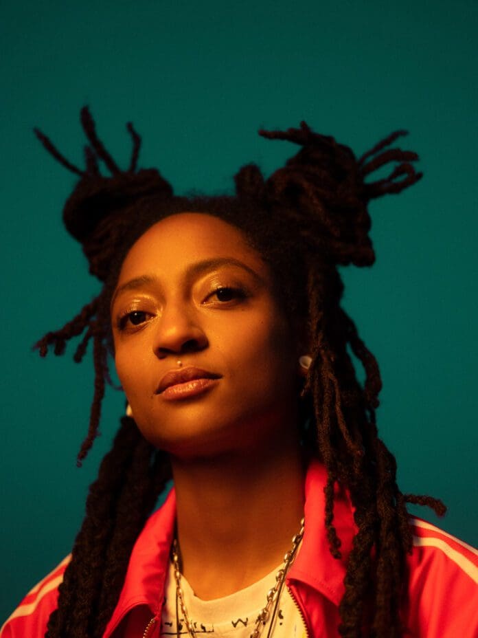 Person with long dreadlocks styled in two buns, wearing a pink jacket with a button over a white shirt, looking forward against a teal background, reminiscent of KT Gorique's unique style.