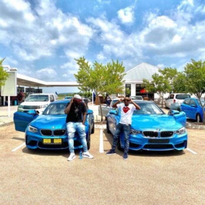 Two individuals stand in front of two identical blue cars in a sunny parking lot. One leans against a car, while the other stands upright. Trees and buildings are visible in the background, creating an atmosphere akin to an eMcimbini music video by Kabza De Small and DJ Maphorisa.