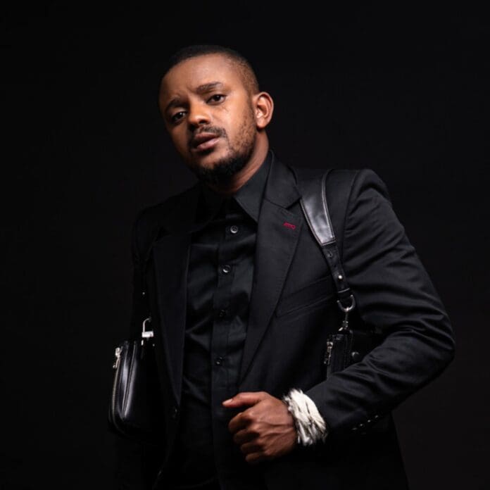 A man wearing a black suit and black shirt poses against a dark background. He has a serious expression, carries a black leather bag with a white rope wrapped around his wrist, and looks like he just left a Konka Amapiano live mix by Kabza De Small.