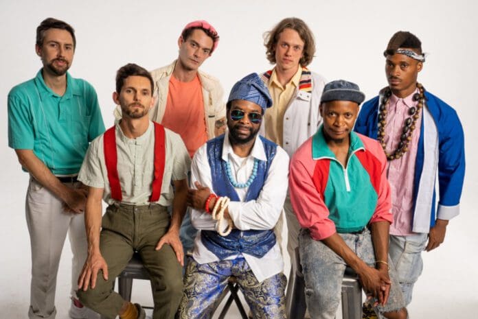 Seven people pose together against a plain white background. They are wearing colorful and varied clothing, reminiscent of an Afrobeat ensemble, with some standing and others seated.