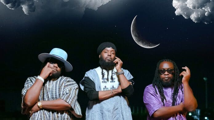 Three men stand confidently under a partly cloudy night sky with a crescent moon. They wear distinctive outfits, including hats, patterned shirts, and sunglasses. As if posing for the cover of their newest 2023 single, they channel the timeless cool of Morgan Heritage.