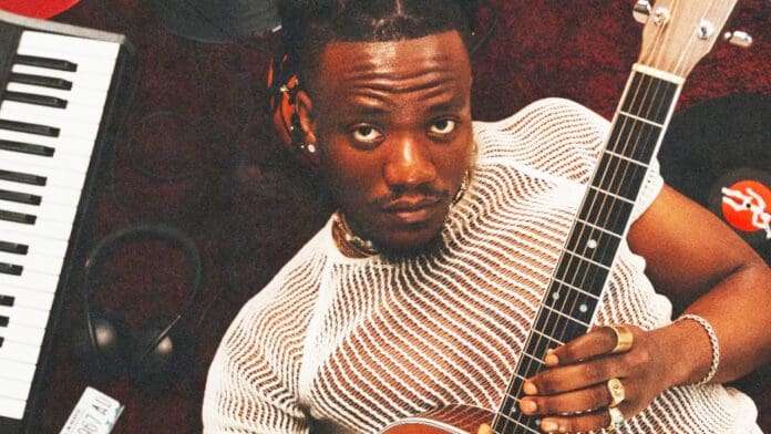 A person in a white mesh shirt holding a guitar, with a keyboard and headphones nearby, looking ready to create the next hit from their debut EP.