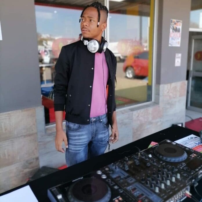 A person wearing NYL headphones stands behind a DJ mixer outside a building, looking off to the side.