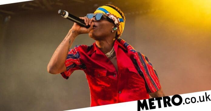 A performer in a red and blue outfit sings into a microphone on stage under vibrant lighting during the London show at Tottenham Hotspur Stadium. The photo has a 