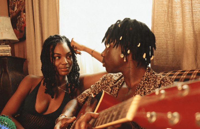 Two people sit on a couch with a guitar, one playing it while the other looks on. Sunlight filters through the window behind them, creating a warm atmosphere in the room, reminiscent of Rema's provocative visuals that often captivate audiences.