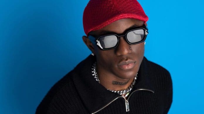 A person wearing a red cap, dark sunglasses with a reflective lens, a black sweater, and a chain necklace, posed against a blue background, looks like Nigerian musician Victony gearing up for his final surgery.