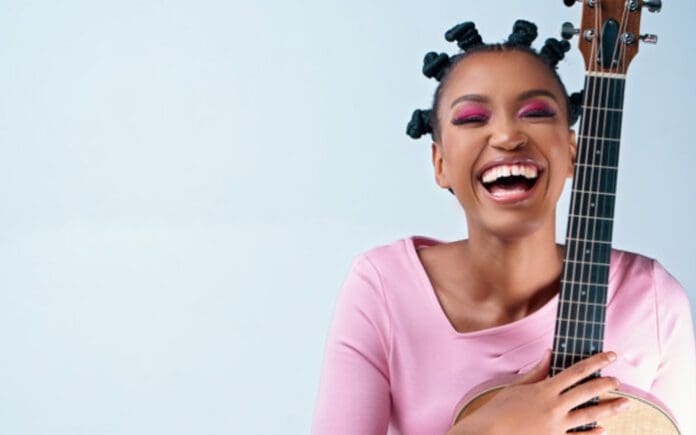 Person with short, twisted hairstyle, in a pink shirt, smiling broadly and holding an acoustic guitar against a light background, preparing for the Phinda Mzala concert.