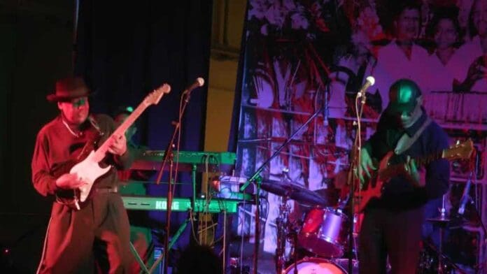 Two musicians perform on stage at the Walnut Street Revue in Louisville, one playing electric guitar and the other on bass guitar, with musical equipment and a colorful backdrop behind them.