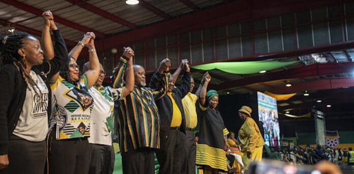 A group of people on a stage hold hands in the air. Some are wearing matching political party attire, symbolizing their commitment to nation building. A large screen displaying images and text is visible in the background.