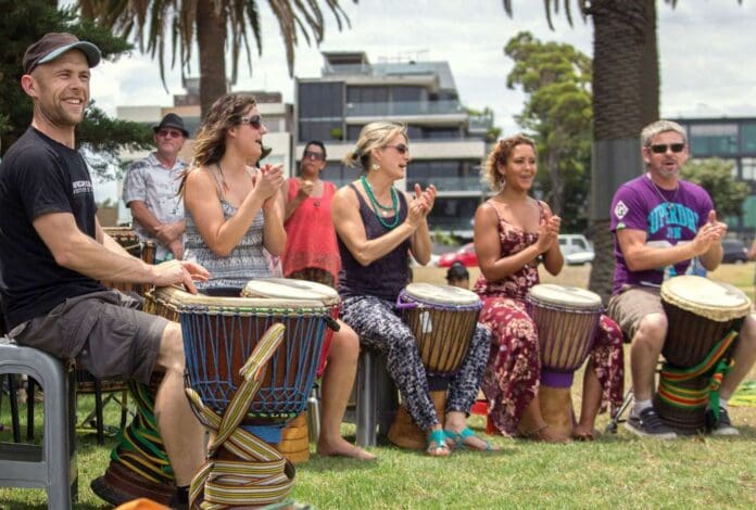 A group of people sitting on chairs outdoors play drums and clap their hands during a lively weekend event in Shepparton. They are surrounded by trees with modern buildings in the background. This vibrant scene is surely making news for its communal spirit and energy.