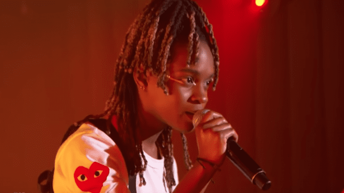 A person with braided hair performs passionately, holding a microphone close to their mouth, under red lighting at the Afro-Carib Festival in the City of Miramar. They are wearing a white shirt with a red heart logo on the sleeve, echoing the vibrant energy of Koffee’s memorable performances.