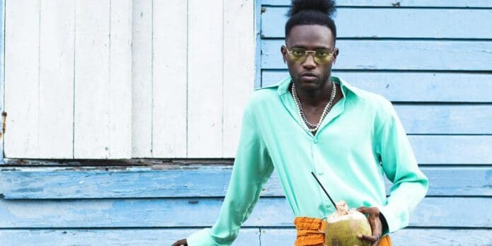 A person wearing a turquoise shirt, orange pants, and glasses stands in front of a blue wooden building, holding a coconut with a straw. It's like something you'd see on OkayAfrica in 2023, celebrating the vibrant style of Nigerian producers.