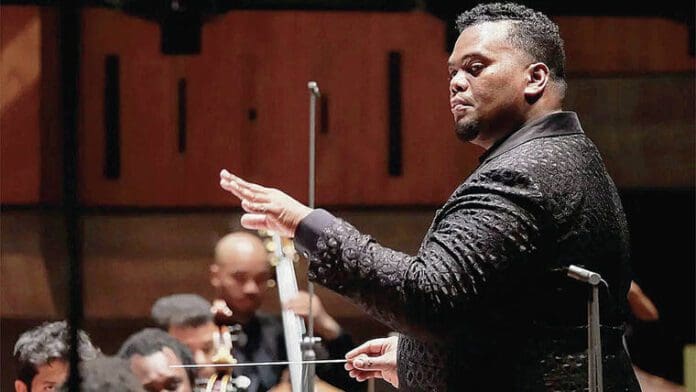 A conductor in a dark patterned suit is leading an orchestra through a classical music performance. The conductor holds a baton and has his left hand raised, guiding the musicians with various instruments in the background. This captivating performance is taking place in February in Honolulu.