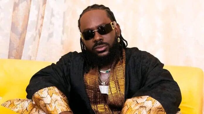 A person wearing sunglasses, a black and gold robe, and jewelry sits on a yellow couch against a light-colored curtain backdrop, reminiscent of an Adekunle Gold recording session with Def Jam vibes.