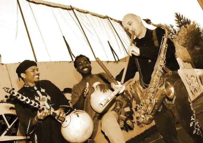 Three men playing saxophones and drums in front of a tent, showcasing the fusion of African music and jazz in a benefit concert at the town church.