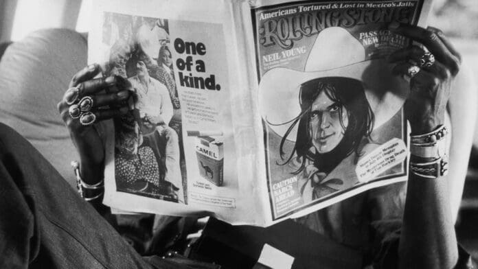 A man is reading a newspaper while sitting on a plane, captivated by Breaking Through Rock Journalism's Boys' Club headline from Rolling Stone.