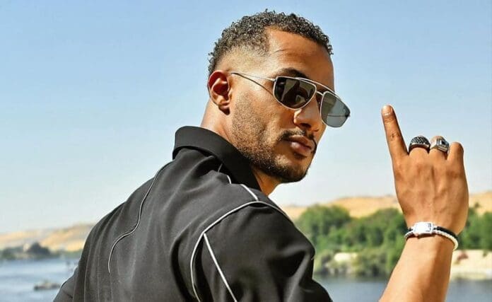  Description: Mohamed Ramadan, an official at the African Cup of Nations, is pointing his finger while wearing sunglasses and a black shirt.