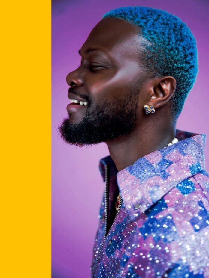 An EP artist with blue hair is smiling in front of a purple background.